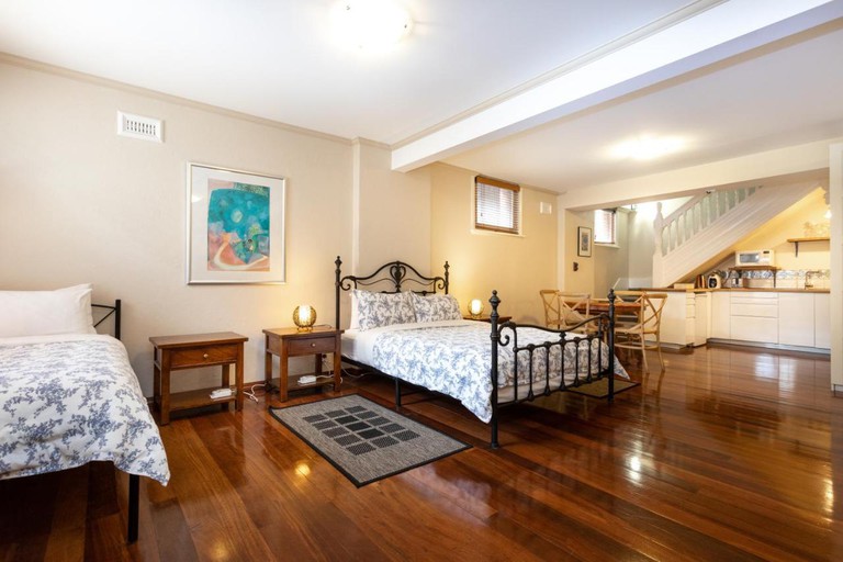 Two double beds stand on the hardwood flooring of a vast, open-plan Fremantle Bed & Breakfast room; on the extreme right, a well-appointed kitchen area sits beneath a staircase