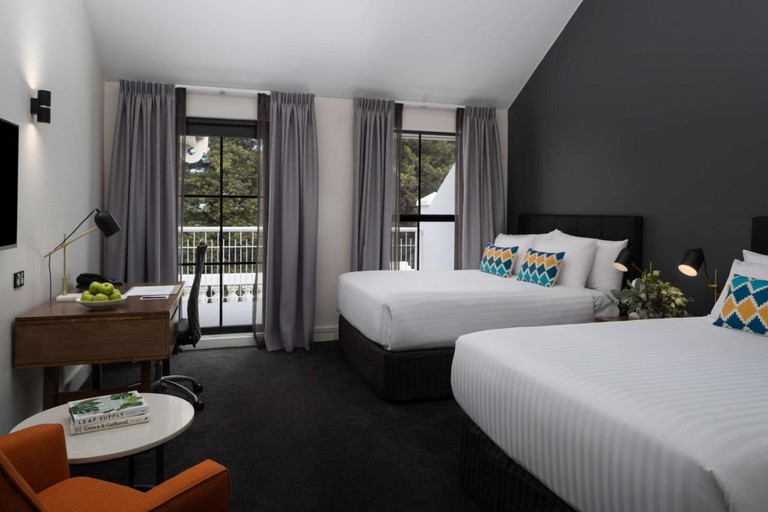 Two double beds in an Esplanade Hotel Fremantle room that features a wall-mounted flat-screen TV with speakers, various lamps, a desk with office chair and apple-filled fruit bowl, a small table and soft chair, and two floor-to-ceiling windows with undrawn curtains