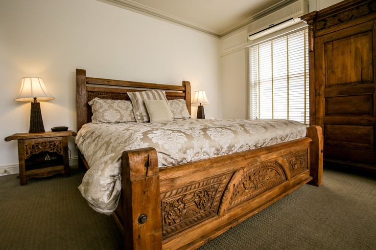 A large, wooden bed in an Allawah Bendigo room features ornate carvings and cream-and-brown covers and pillows, matching the wider decor; a bedside table and lamp, overhead air conditioning, a large wooden cabinet and undrawn window blinds can also be seen