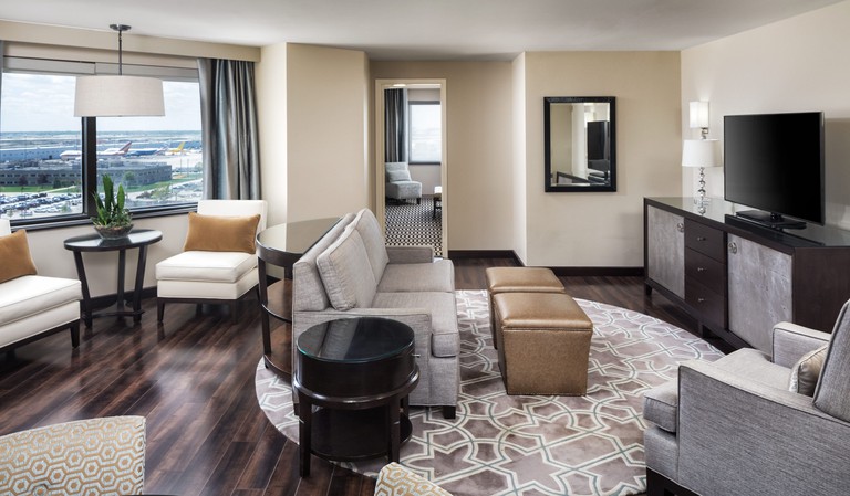 A cream-toned lounge area in a suite at Sheraton Suites Chicago O'Hare with a gray couch facing a TV, dark hardwood floors and various other seating