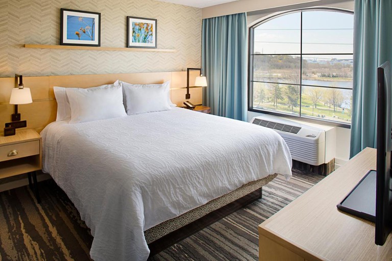 A hotel room at Hilton Garden Inn with a grey striped carpet, light blue curtains, a white bed and two floral pictures on the back wall