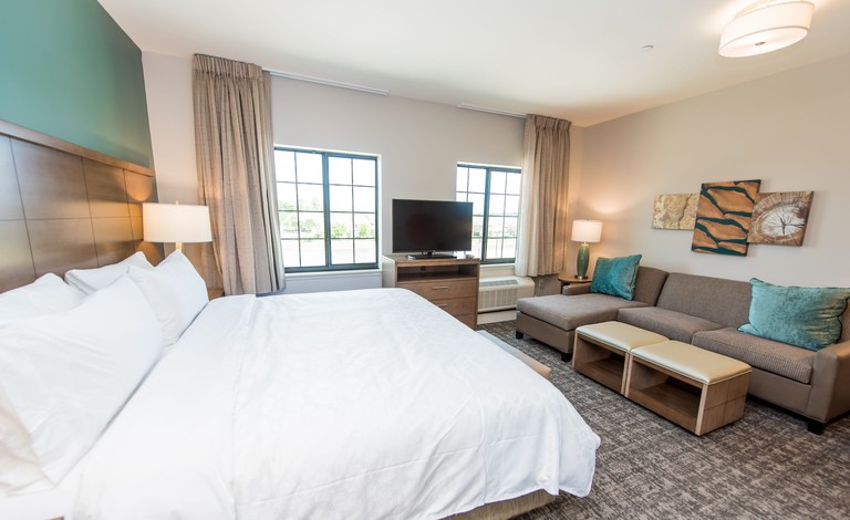 A king bed studio suite with a sofa and art opposite the bed and a dresser with a TV between two windows at the Staybridge Suites Houston – Humble Beltway 8 E