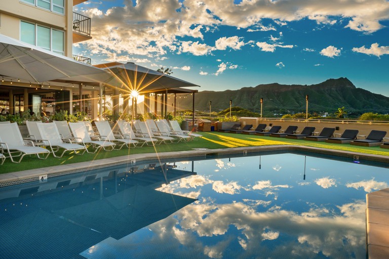 Queen Kapiolani Hotel outdoor pool with loungers, mountains in the distance and a bar beside it at sunrise