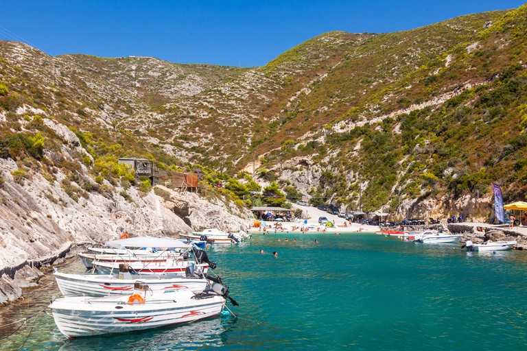 Sea view of Porto Vromi Anafonitria, a small rocky inlet sheltered by scrub-covered hills, with motorboats moored at either side and a small white-sand beach.
