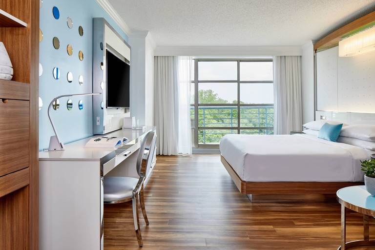 A modern bedroom at Renaissance Charlotte SouthPark with a double bed, hardwood floors, a baby blue accent wall, a white writing desk, a TV and a private balcony