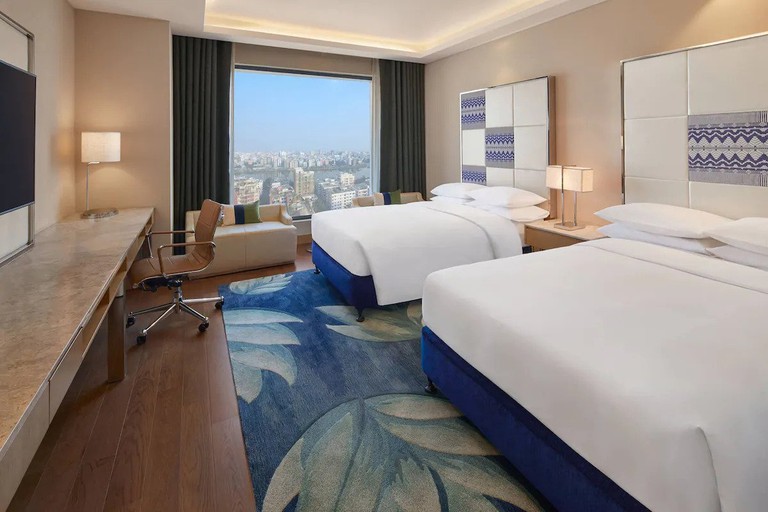 A guest room with two twin beds, a bold area rug, seating by the windows and city views at the Renaissance Dhaka Gulshan Hotel