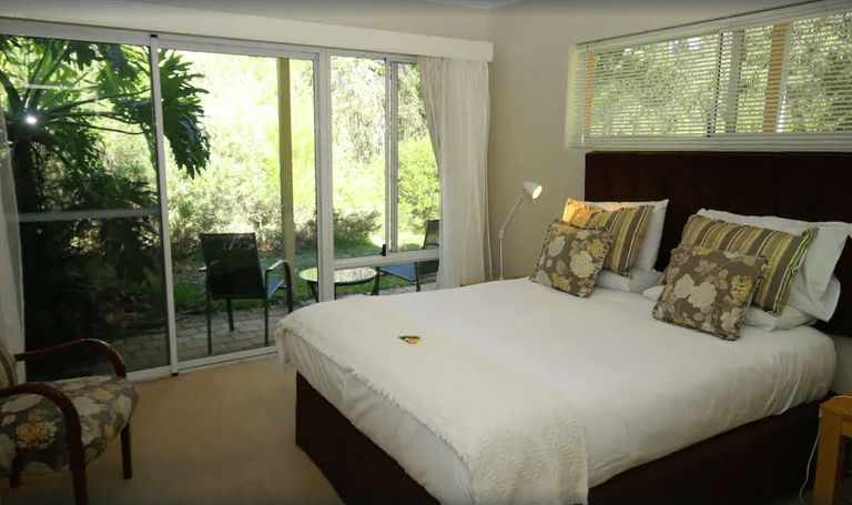 A cosy guest room with a bed topped with floral and striped pillows and a small furnished veranda with forest views