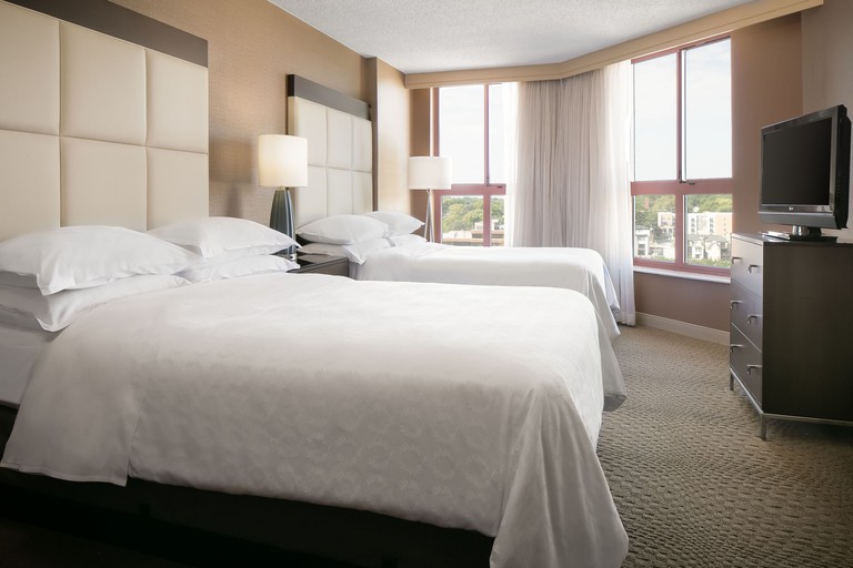 A guest room with two double beds, windows with city views and a dresser topped with a TV at the Sheraton Suites Country Club Plaza