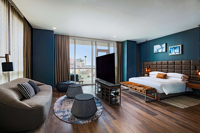 Modern-looking bedroom with cobalt blue walls, a large bed, a TV in the middle of the room, a spacious couch and a private balcony at Thompson San Antonio – Riverwalk