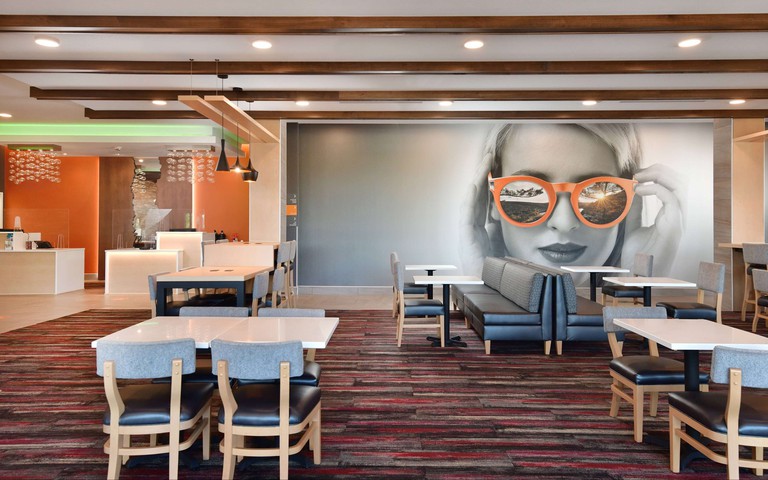 Dining area at La Quinta Inn & Suites by Wyndham, with black-and-red striped carpet, white tables, black leather seating and large wall mural of a woman wearing orange sunglasses.