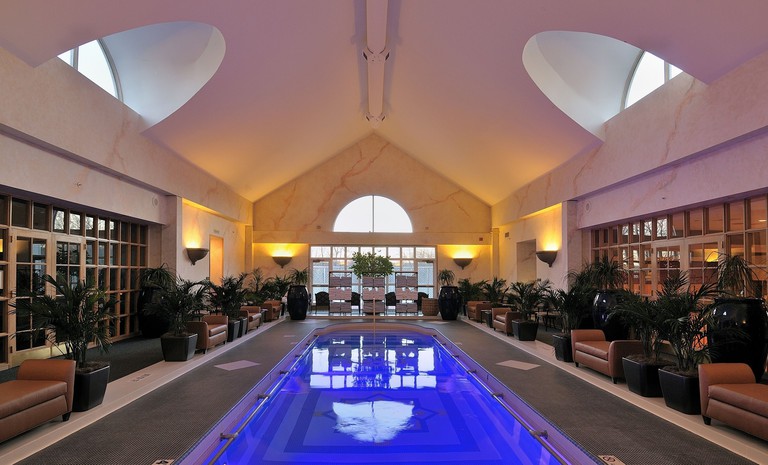 Electric blue indoor pool lined with potted palms and leather benches at the Spa at Norwich Inn