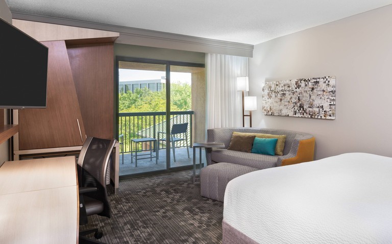 Double room at Courtyard by Marriott Lafayette Airport, with white bed, gray sofa and carpet, and floor-to-ceiling window that opens onto a small railed balcony.
