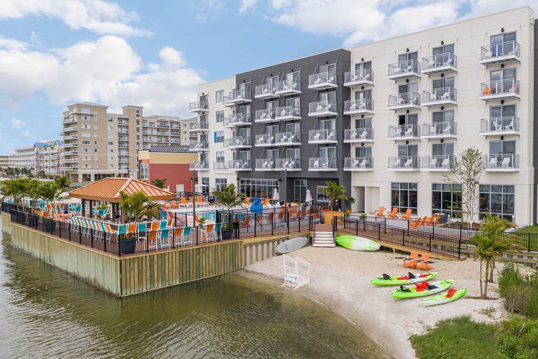 Waterfront area at Aloft Ocean City, with a small sandy section housing green kayaks and an outdoor pool, both overlooked by a modern gray and white building with balconied rooms.