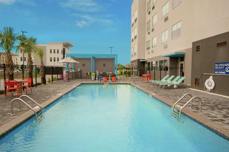 Large fenced-off rectangular outdoor pool at Hilton Lafayette River Ranch, overlooked by a tall beige building and surrounded by turquoise sun-loungers and pink chairs.