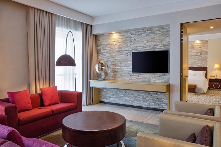 Separate living area in a bedroom at the Westin City Centre Bahrain with two red couches, two cream leather chairs, a brown coffee table, a brick wall and a flat screen TV