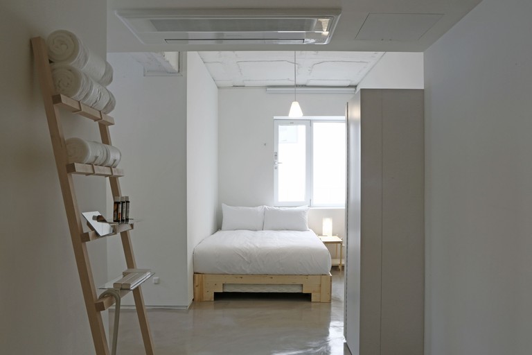 A light bedroom at Small House Big Door, with white walls, a cushioned bed next to windows, a bedside table and lamp