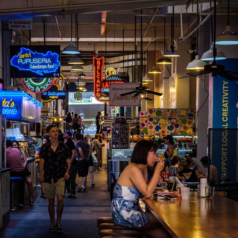Tourists and locals peruse the restaurant outlets at Grand Central Market with neon signs overhead