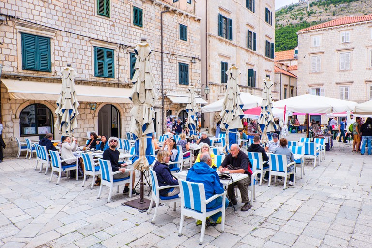 Diners sitting on blue-and-white striped chairs at Kamenice's outdoor tables in a stone-paved square in the old town