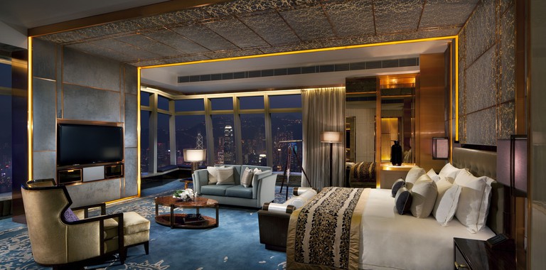 A stylish room with a cushioned bed, a TV, a blue carpet, a light-blue sofa, an armchair and city views at The Ritz Carlton Hong Kong