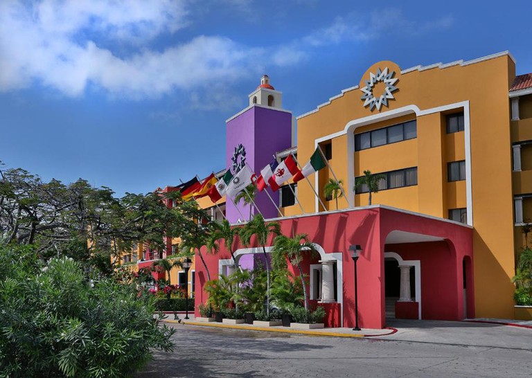 The colorful red, purple and ocher exterior of Hotel Adhara Cancún, with flags of different countries flying from the porte-cochère
