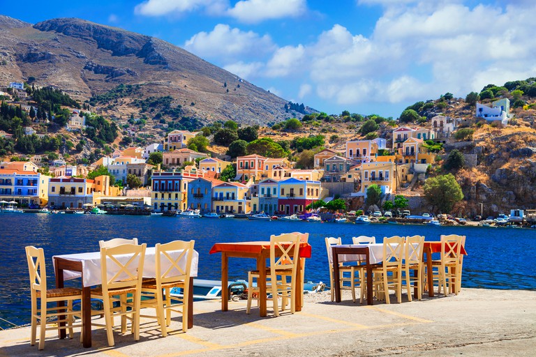 Waterfront dining tables on Symi Island, with colourful buildings and tree-speckled hills in the background